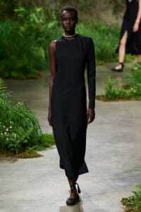 Model walking a garden runway in a black, one-shoulder ankle-length dress with a pearl choker and black lace-up sandals