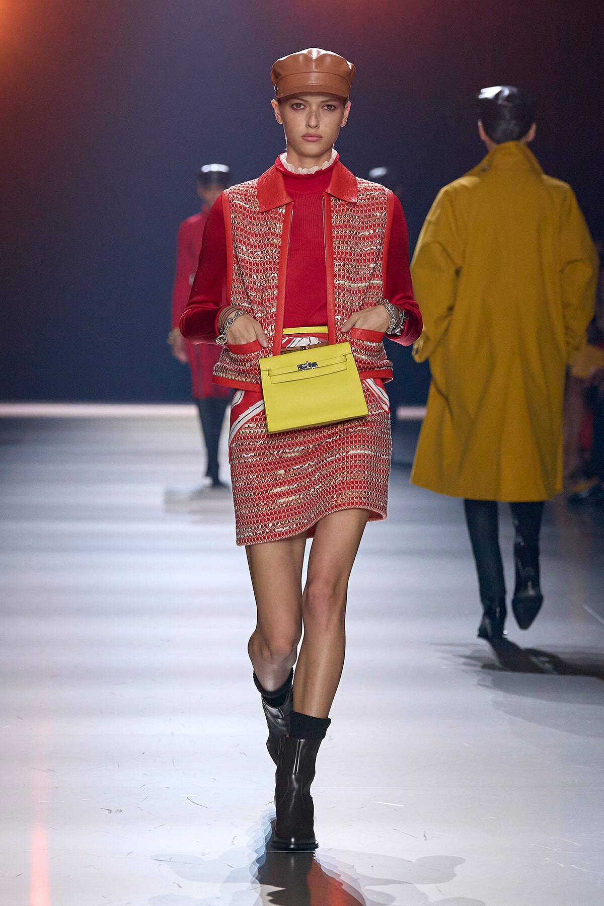 Model on a fashion runway wearing a red tweed vest and matching red skirt with a red top, brown leather cap, and dark ankle boots, carrying a bright yellow handbag.