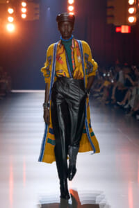 Model walking a fashion runway in a bright yellow patterned cardigan over a green turtleneck, black leather pants, and a black leather cap under bright lights, audience in background.