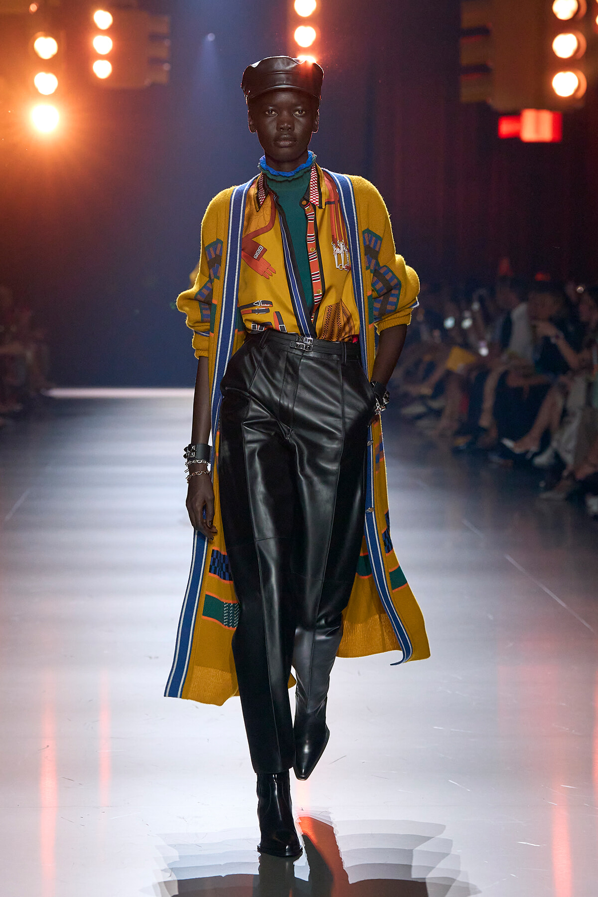 Model walking a fashion runway in a bright yellow patterned cardigan over a green turtleneck, black leather pants, and a black leather cap under bright lights, audience in background.