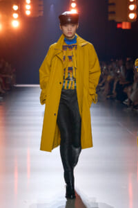 Runway model wearing a mustard-yellow trench over a colorful patterned top, blue scarf, and a black cap, walking the runway in pinstripe pants and black boots.