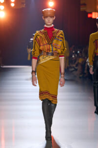 Model walking the runway in a mustard yellow knit dress with red striped blouse, wide belt, and knee-high black boots; orange cap and bold military-inspired detailing in warm tones.