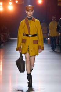Model walking the runway in a mustard yellow trench coat with brown accents, brown cap, and a black bag at a fashion show.