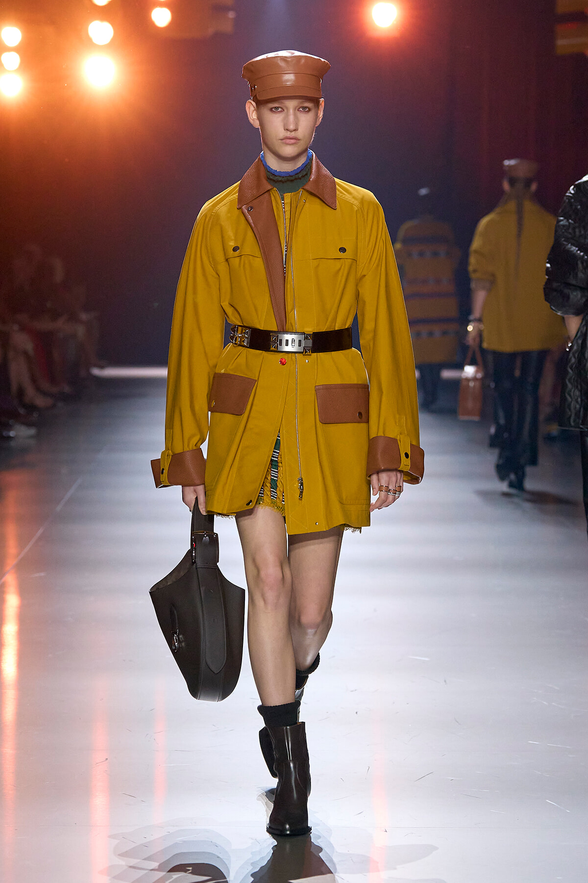 Model walking the runway in a mustard yellow trench coat with brown accents, brown cap, and a black bag at a fashion show.