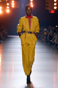 Model walks the runway in a mustard yellow jumpsuit with a wide brown belt, red turtleneck, and black ankle boots.