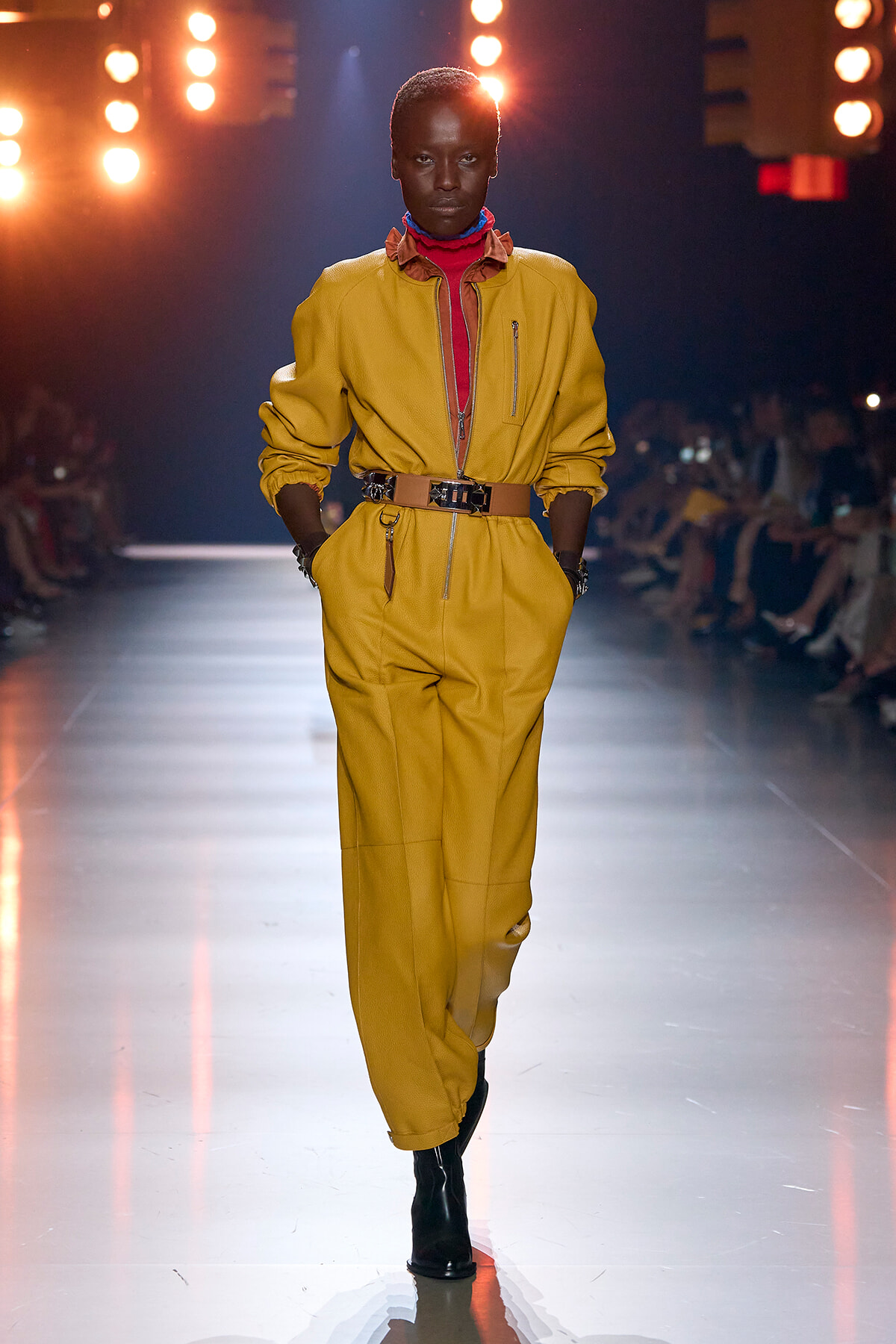 Model walks the runway in a mustard yellow jumpsuit with a wide brown belt, red turtleneck, and black ankle boots.