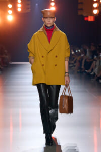 Model on a runway wearing a mustard oversized double-breasted coat with rolled sleeves, red turtleneck, black leather pants, brown cap, and a brown structured handbag.