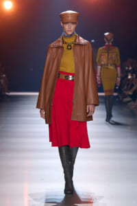 Model walking the runway in a brown leather cape over a mustard sweater and red midi skirt, paired with black knee-high boots and a matching brown cap.