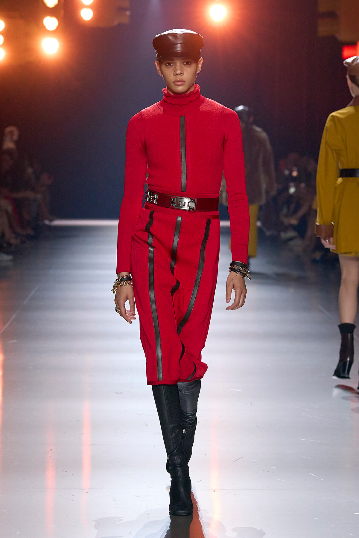 Model walking a fashion runway in a red turtleneck jumpsuit with black vertical stripes, a wide metallic belt, knee-high black boots, and a black leather cap.