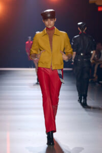 Model walks the runway in a mustard yellow zip-front jacket, red pants, and a black cap. Hands in pockets.