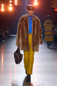 Model walks runway in oversized brown fur coat, bright blue turtleneck, mustard-yellow pants, black cap, carrying a dark brown handbag.