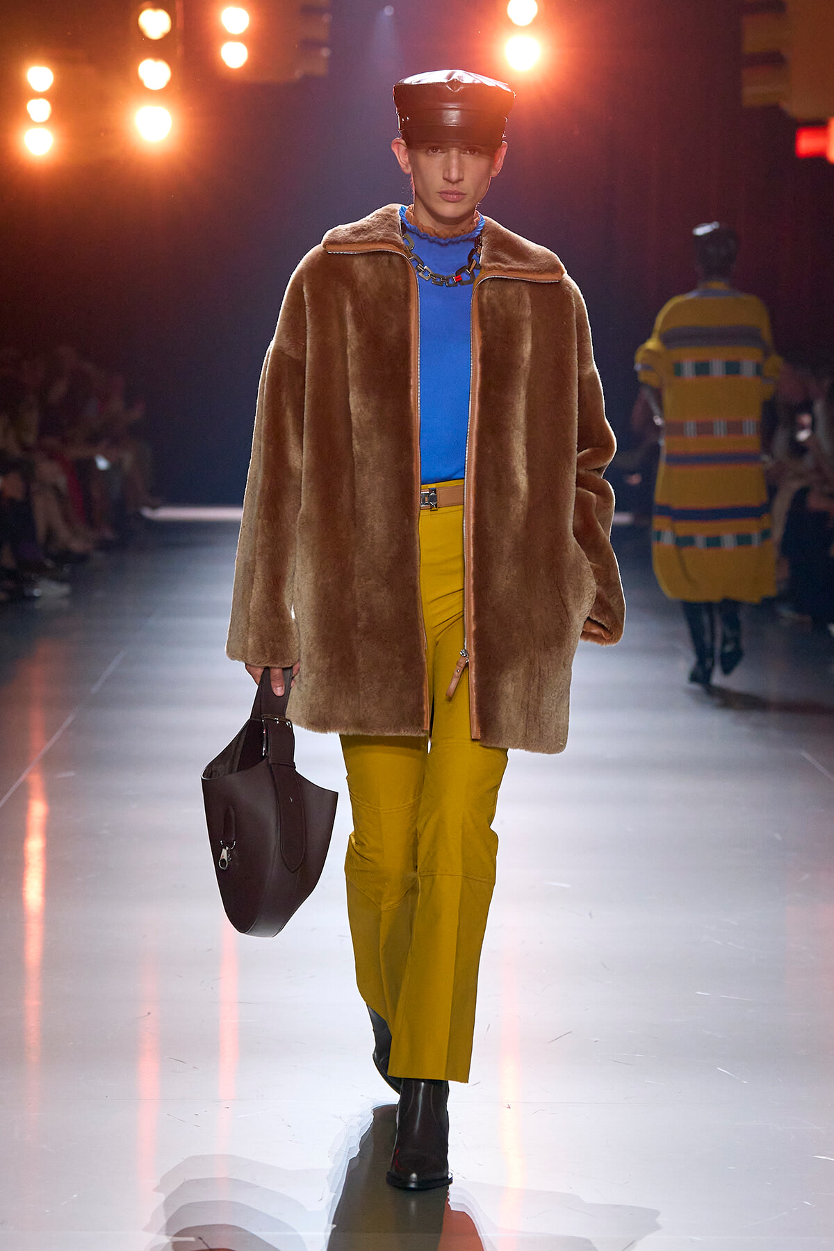 Model walks runway in oversized brown fur coat, bright blue turtleneck, mustard-yellow pants, black cap, carrying a dark brown handbag.