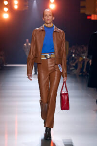 Model walks a fashion runway wearing a brown leather suit with a blue top, beige belt, and a red handbag in hand.