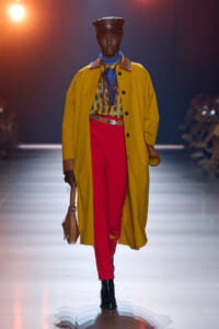 Model walks the runway in a mustard trench over a striped blouse and red trousers, accessorized with a blue neck scarf, black cap, and tan handbag.