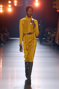 Model on a runway wearing a mustard-yellow matching jacket and pencil skirt with a brown belt and black knee-high boots.