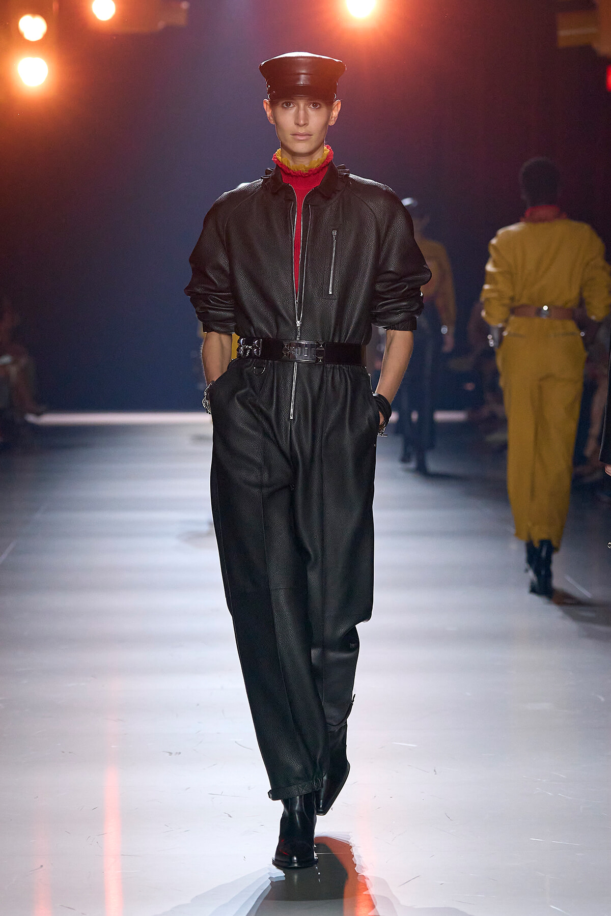 Model walks a fashion runway in a black leather jumpsuit with a red turtleneck, wide belt, and black cap under warm stage lights.