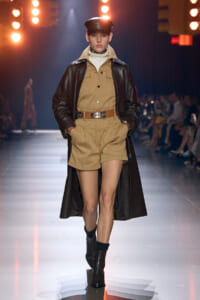 Model on a runway wearing a beige utility romper with a wide brown belt and a long dark leather coat, black boots, and a cap under bright stage lights.