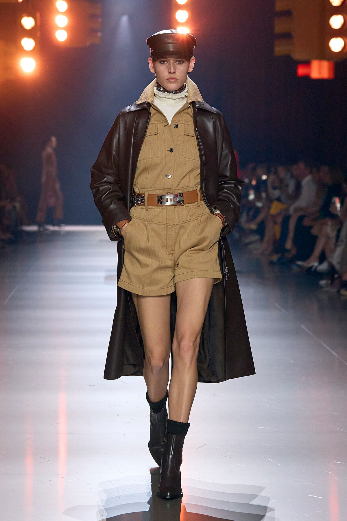Model on a runway wearing a beige utility romper with a wide brown belt and a long dark leather coat, black boots, and a cap under bright stage lights.