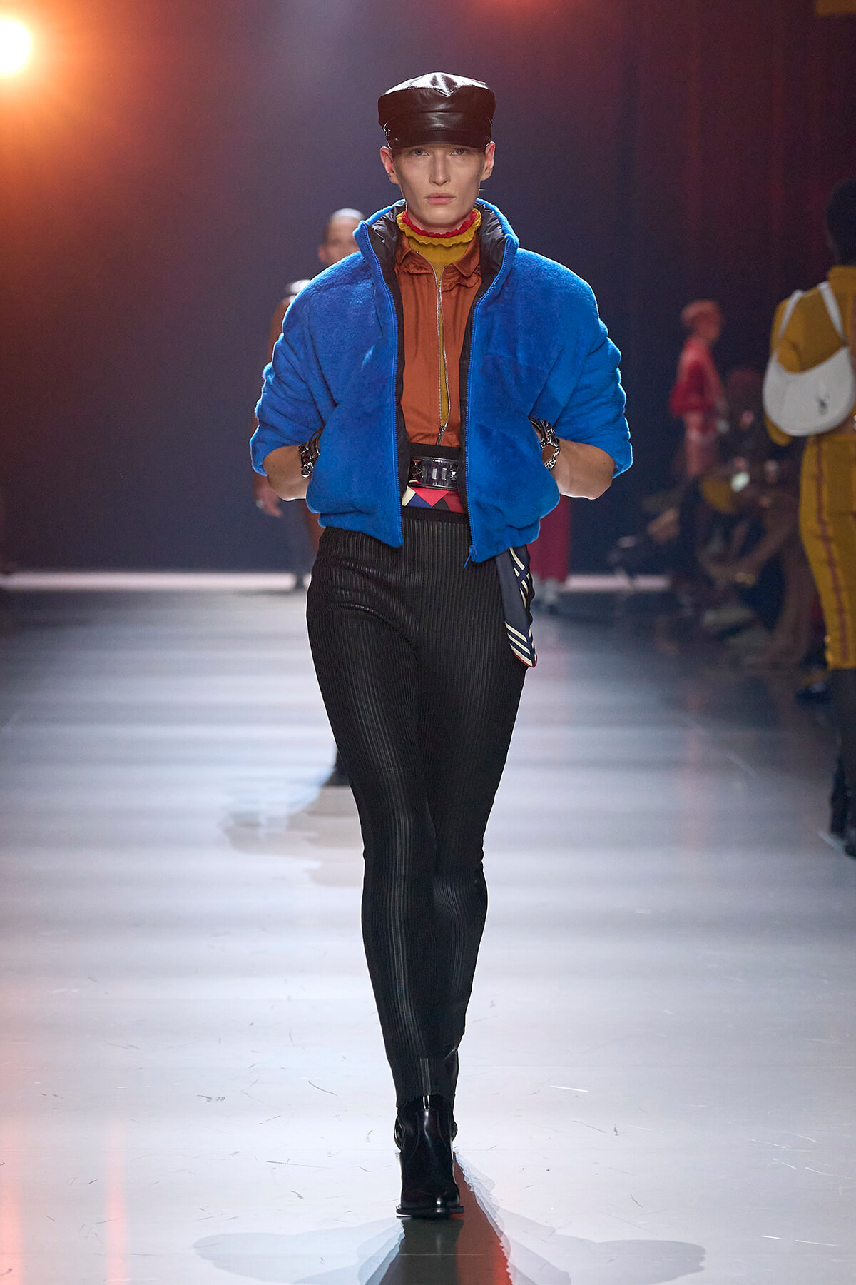Model on a runway wearing a bright blue puffer jacket over an orange-brown shirt, black pinstripe pants, a black cap, and black boots.