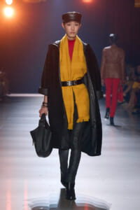 Model on a fashion runway wearing a mustard-yellow coat layered with a black outer coat, a bright yellow scarf, black belt, and a black handbag.
