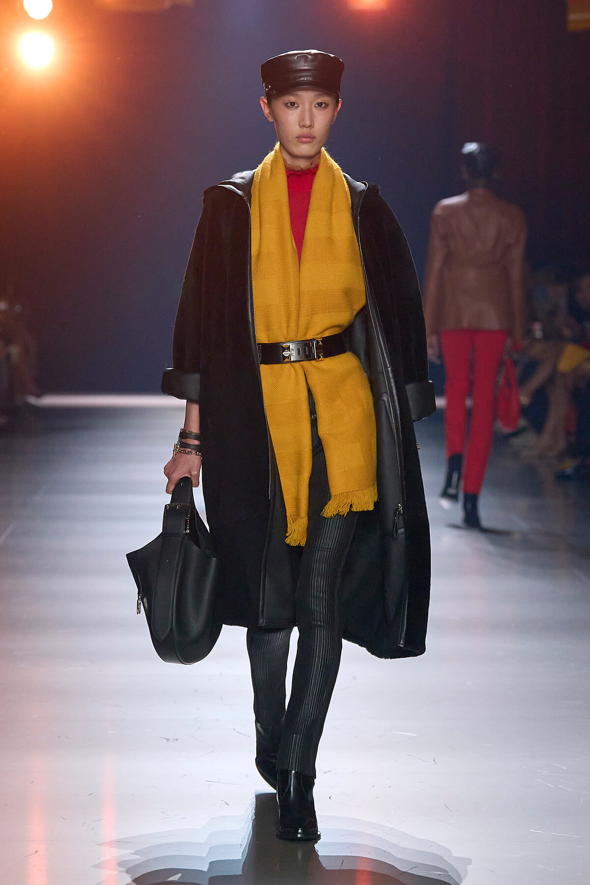 Model on a fashion runway wearing a mustard-yellow coat layered with a black outer coat, a bright yellow scarf, black belt, and a black handbag.