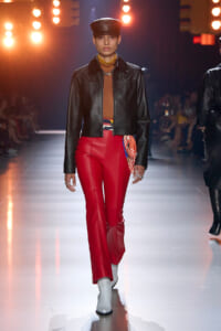 Model walks a fashion show wearing a black leather jacket, camel top, and bright red leather pants with white boots and a black cap.