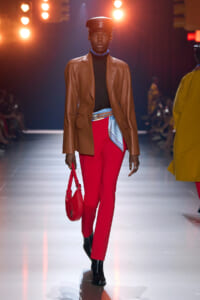 Model walking the runway in a brown leather blazer, black turtleneck, and bright red pants, with a blue scarf-like belt, red handbag, and black ankle boots under warm stage lights