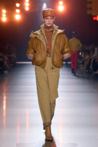 Model walks the runway in camel-tan tones: oversized brown leather puffer jacket, matching tan trousers, and brown cap with boots.