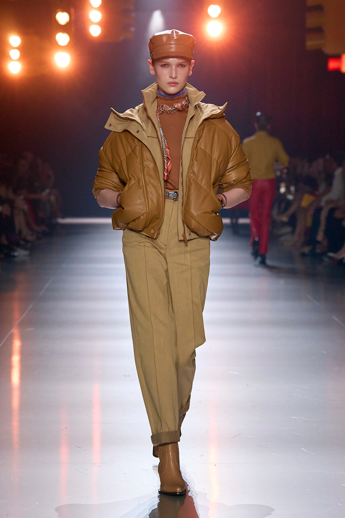 Model walks the runway in camel-tan tones: oversized brown leather puffer jacket, matching tan trousers, and brown cap with boots.