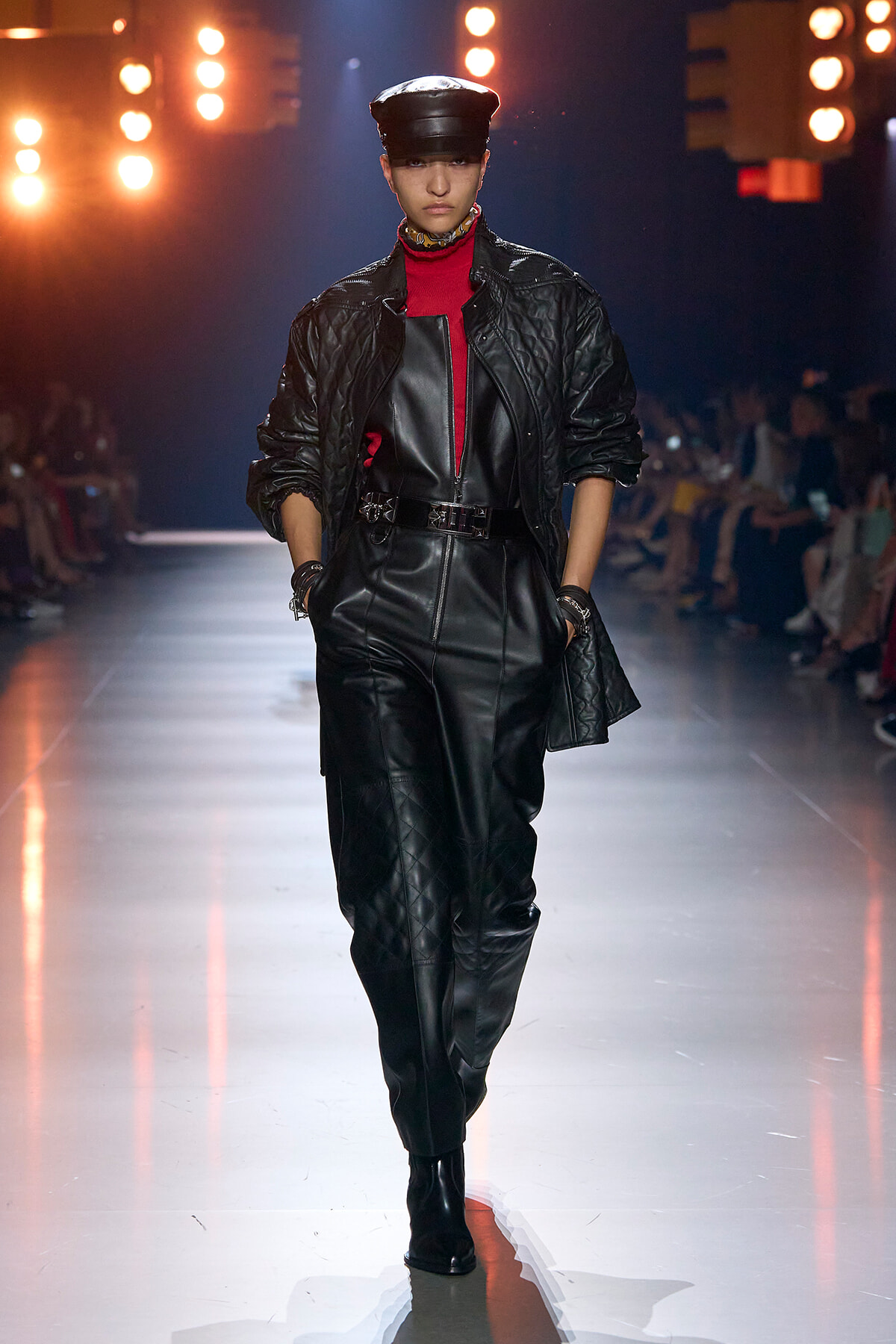 Model wearing a black quilted jacket over a red turtleneck and belted leather jumpsuit on a fashion runway with audience in the background in soft focus.