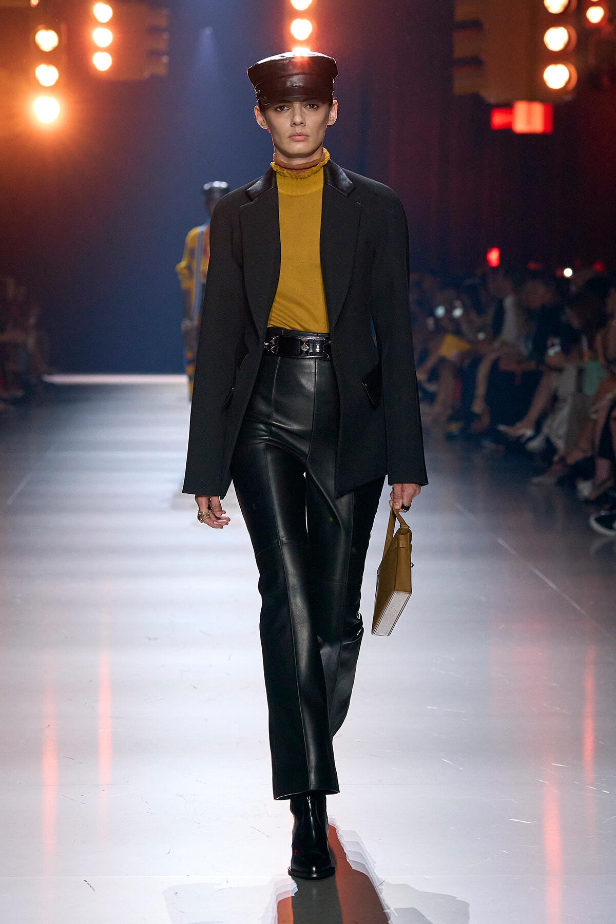 Model on a fashion runway wearing a mustard turtleneck with a black blazer, black leather pants, black ankle boots, a black hat, and carrying a tan clutch.