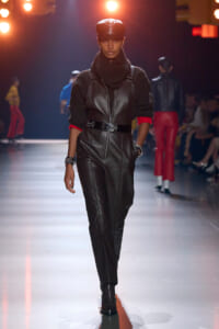 Model walks a fashion show wearing a black leather jumpsuit with a front zipper and belt, layered over a dark knit sweater and cap under warm runway lights.