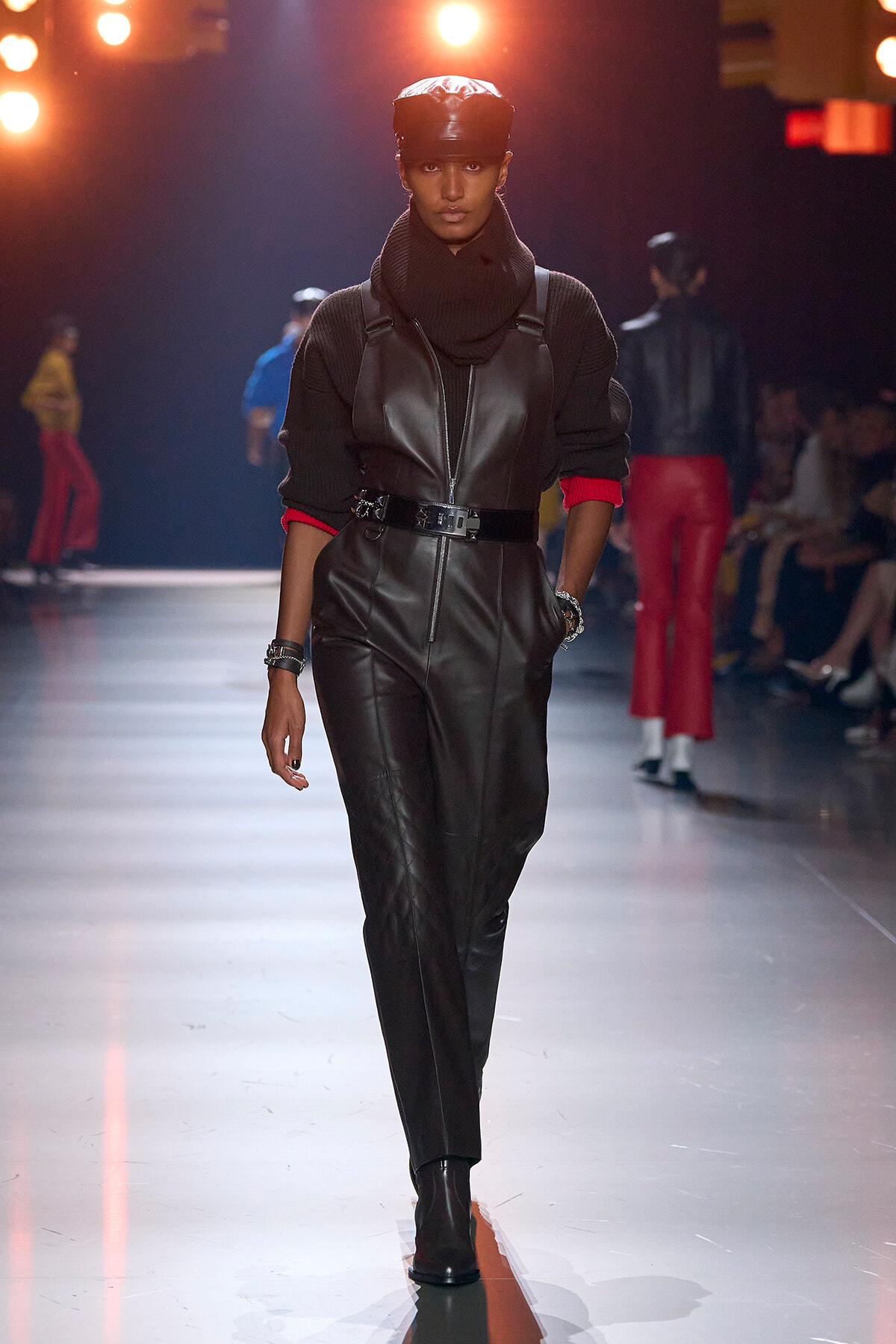 Model walks a fashion show wearing a black leather jumpsuit with a front zipper and belt, layered over a dark knit sweater and cap under warm runway lights.