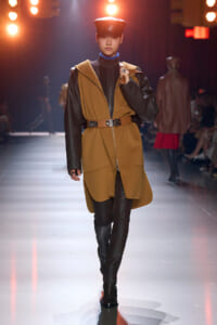 Model walks a fashion show in a camel and black coat with a wide belt, black ribbed pants, and a cap under stage lights.