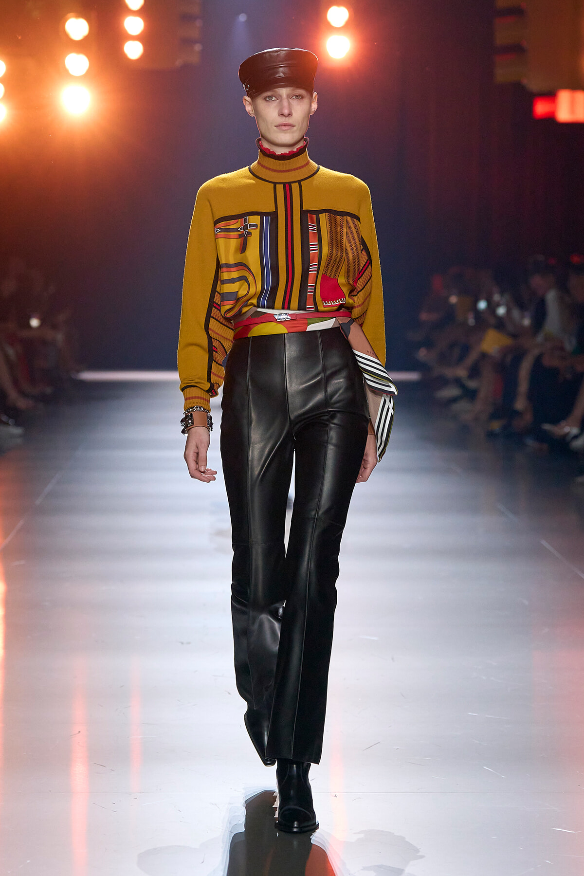 Model walks the runway in a mustard knit top with bold color-block stripes, paired with black leather pants and a black cap.