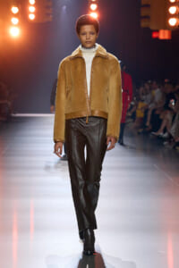 Model walks the runway in a camel suede studded jacket over a white turtleneck, black leather pants, and ankle boots with bright stage lights and an audience in the background