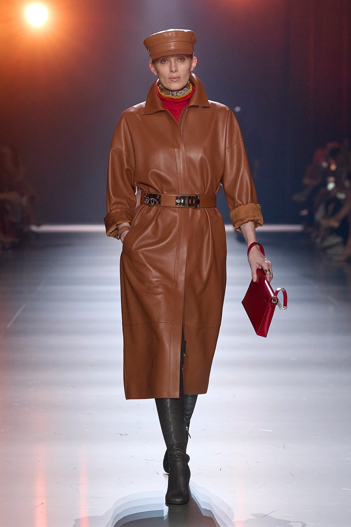 Model walking a runway in a tan leather belted long coat, matching cap, red turtleneck, black boots, and a red handbag.
