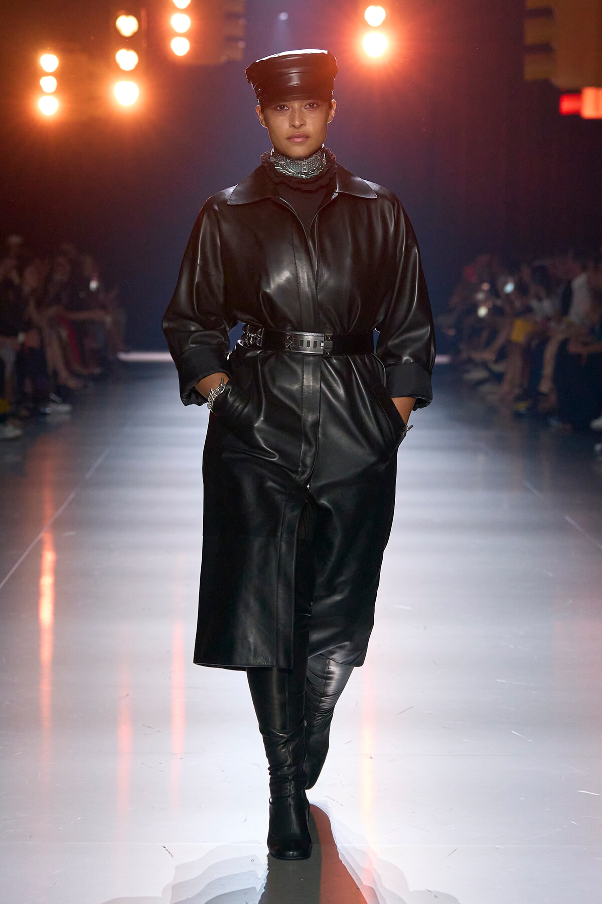 Model walks the runway in a black leather belted coat, matching knee-high boots and a black cap under warm stage lights