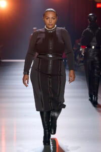 Plus-size model walking a fashion runway in a black, structured dress with front zipper, wide belt, and knee-high black boots, under stage lighting.
