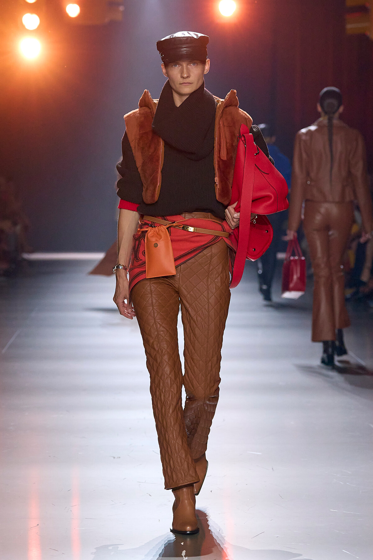 Model walks a fashion runway wearing brown quilted pants, a black sweater, orange vest, tan boots, and carries a red bag with an orange pouch against warm stage lights.