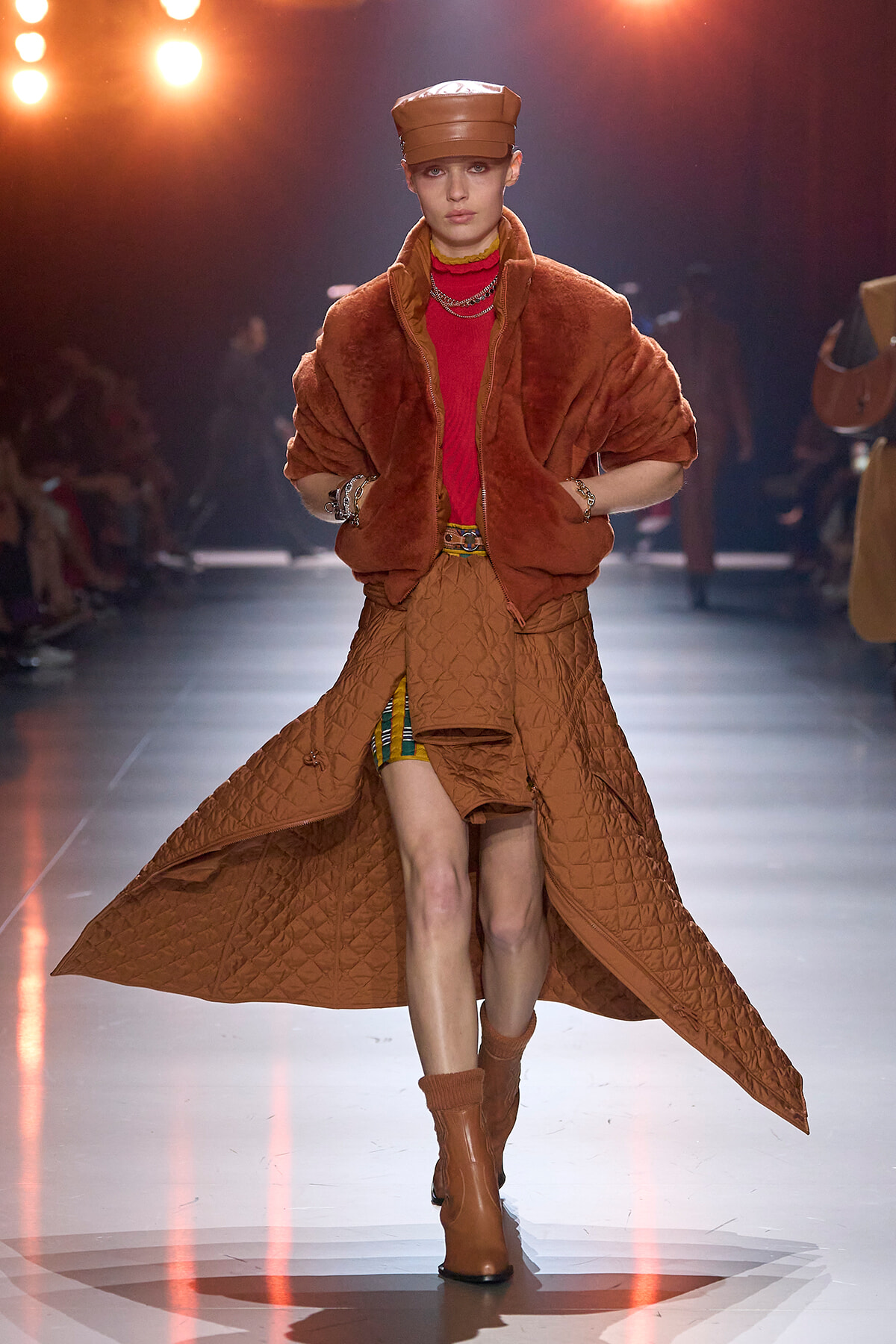 Model walking a fashion runway in a brown quilted long coat over a red top, with a matching brown cap and boots.