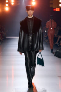 Model walks the runway in a black leather coat with a large fur collar, matching leather pants, and a dark cap, carrying a teal handbag.