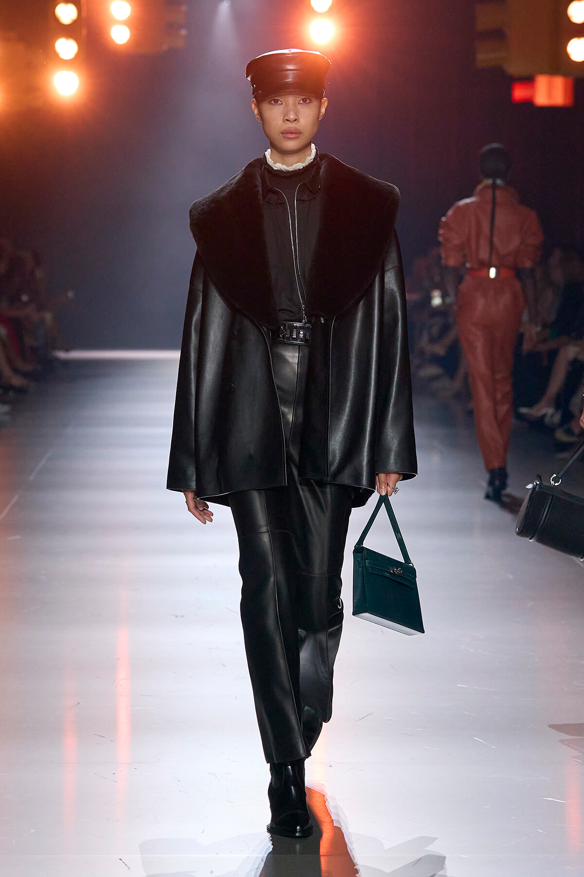 Model walks the runway in a black leather coat with a large fur collar, matching leather pants, and a dark cap, carrying a teal handbag.