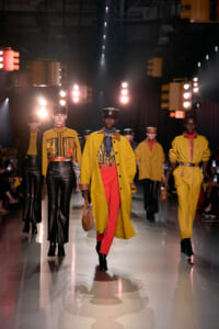 Group of models walking a runway in mustard-yellow coats, red pants, and leather accessories under bright stage lights.