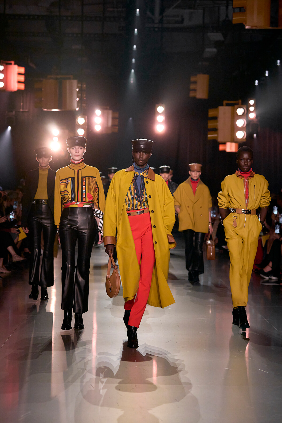 Group of models walking a runway in mustard-yellow coats, red pants, and leather accessories under bright stage lights.
