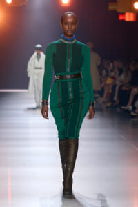 Model struts a runway in an emerald green, form‑fitting dress with a black belt, blue ruffled collar, and knee‑high brown boots.