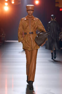 Model walking a runway in a tan leather coat with geometric stitching, matching belt and pants, and a brown cap, carrying a large gray bag mid-stride.