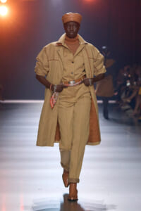 Model walks the runway in a monochrome tan ensemble: trench coat layered over a tan suit with a belt and brown cap, mid-stride under warm lighting.