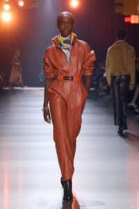 Model walks the runway wearing an orange-brown leather jumpsuit with a matching belt and a colorful scarf, black ankle boots.