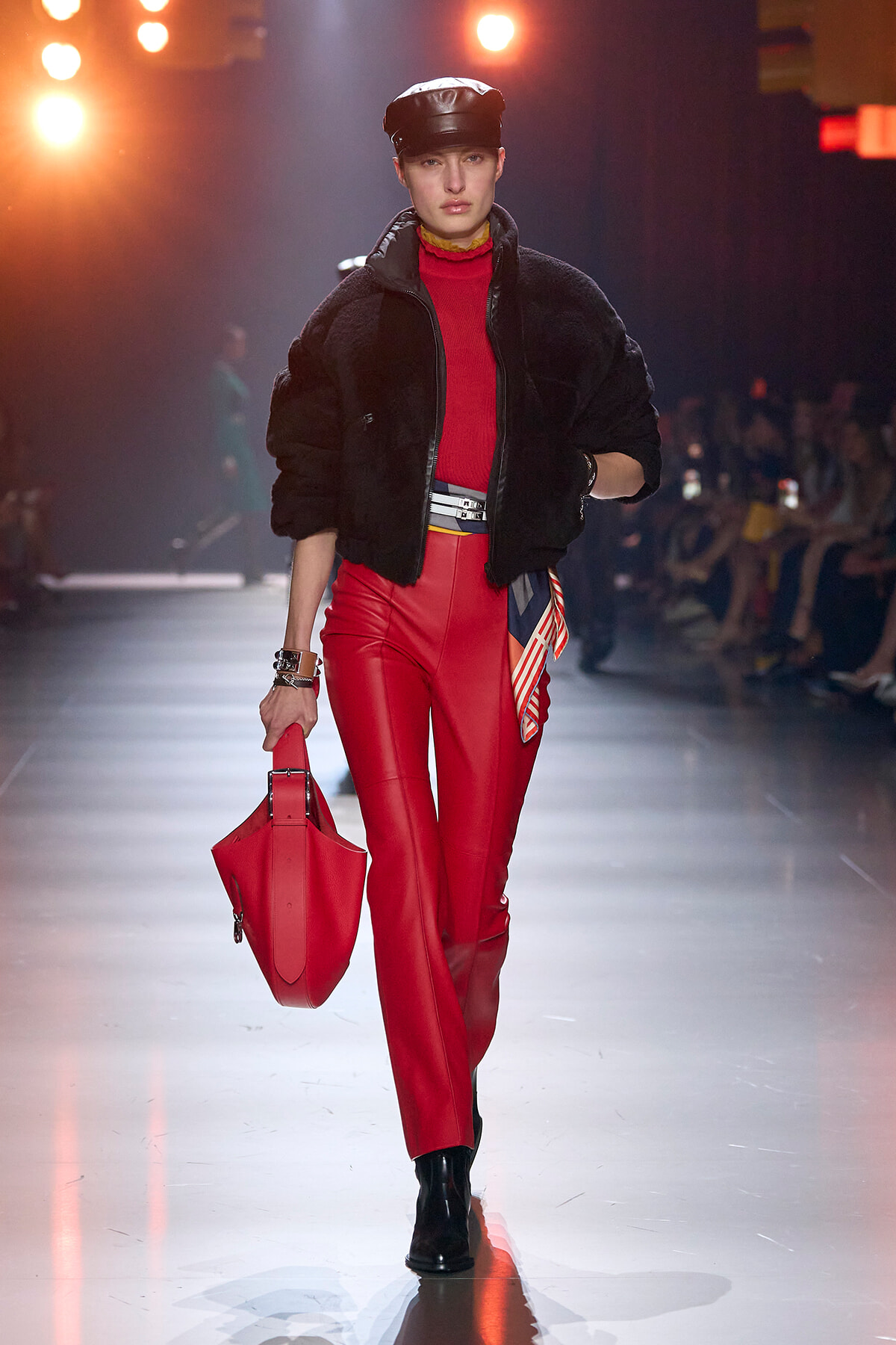 Model walks the runway in red leather pants and a red turtleneck under a black fur jacket, with a black cap and red handbag.